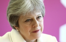 A portrait of British Prime Minister Theresa against a pink background. 
